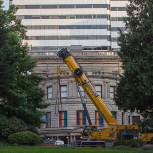 Front image of the Portland City Hall covered by two trees on two end and a yellow crane, windows of the building with rainbow flags