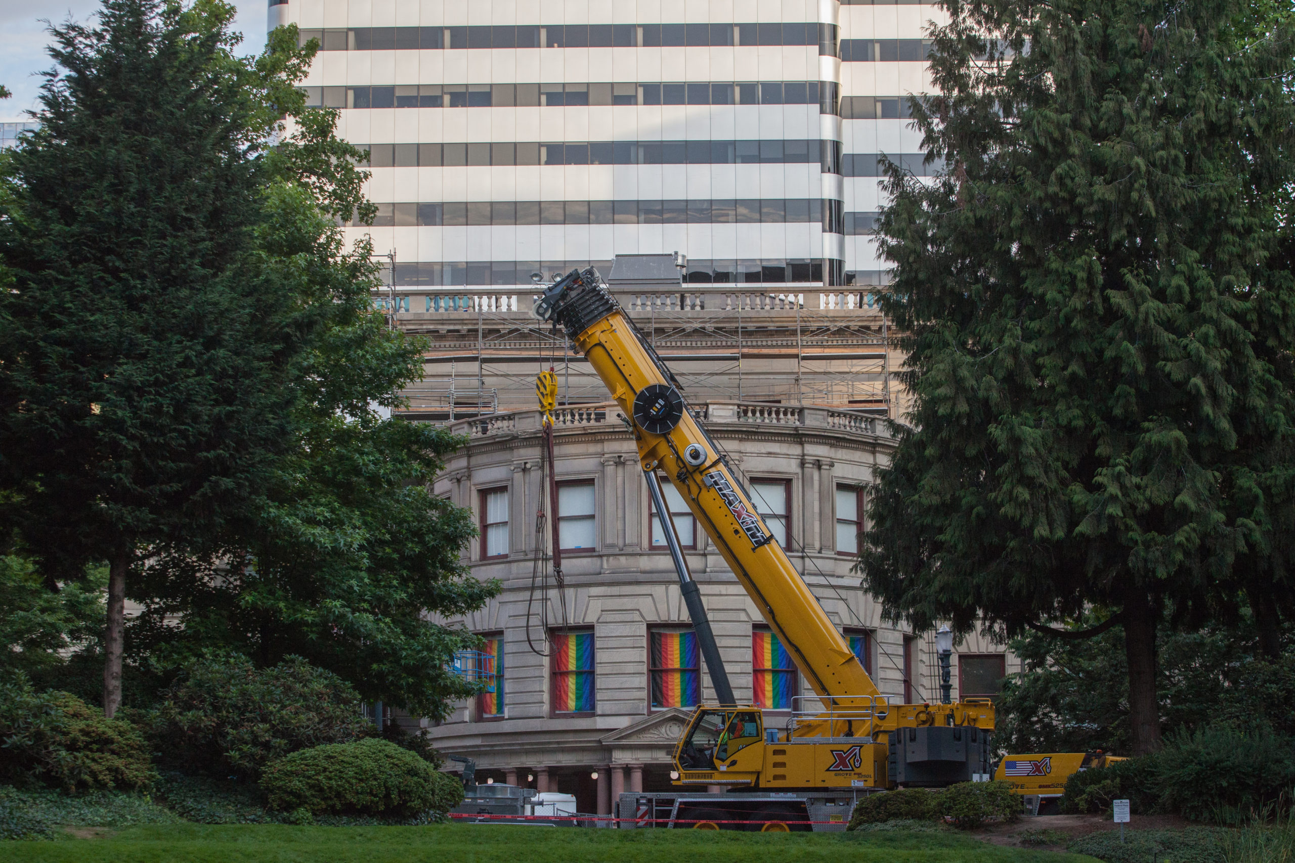 Front image of the Portland City Hall covered by two trees on two end and a yellow crane, windows of the building with rainbow flags